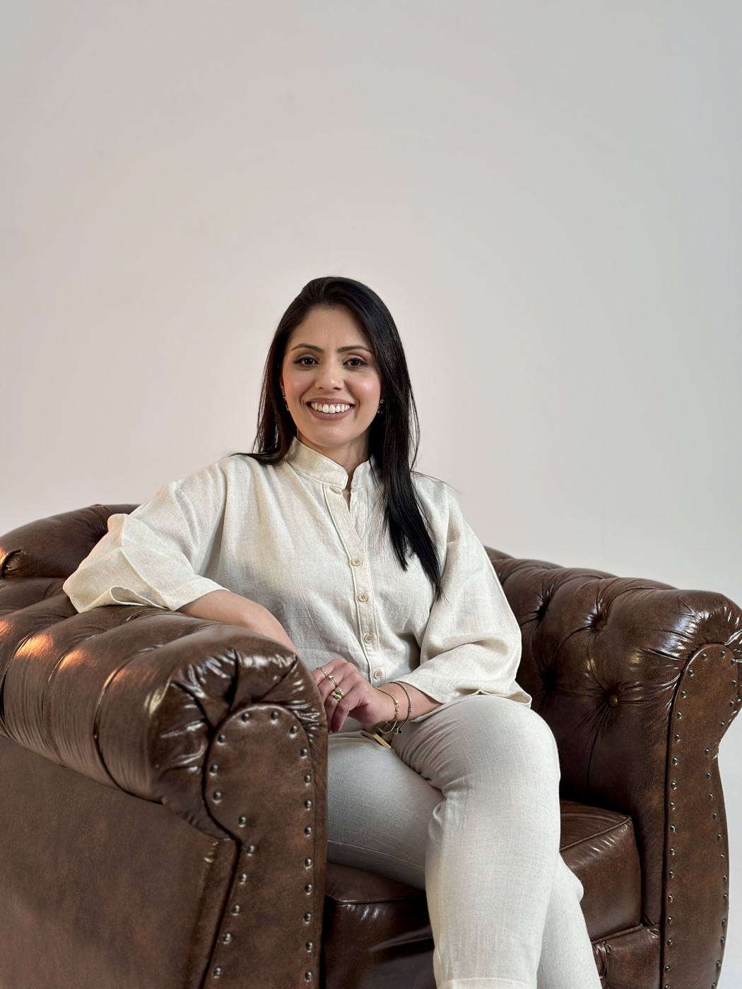 Cássia de Faveri sorrindo, sentada em uma poltrona de couro marrom.